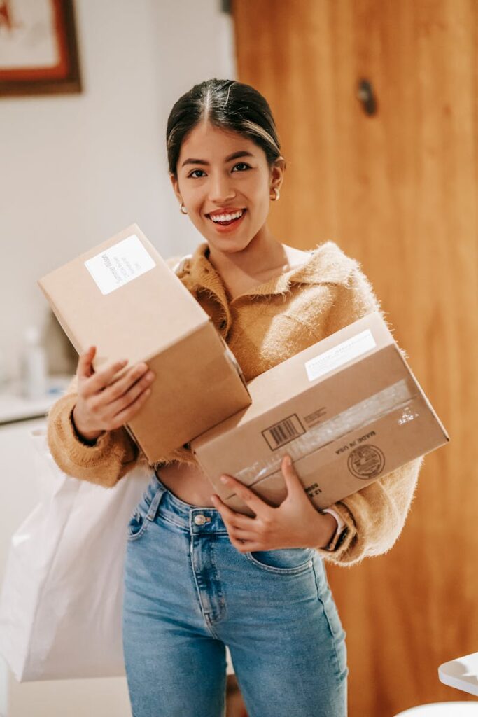 Smiling young ethnic lady in casual outfit standing in light room in house with packages and shopping bag while looking at camera